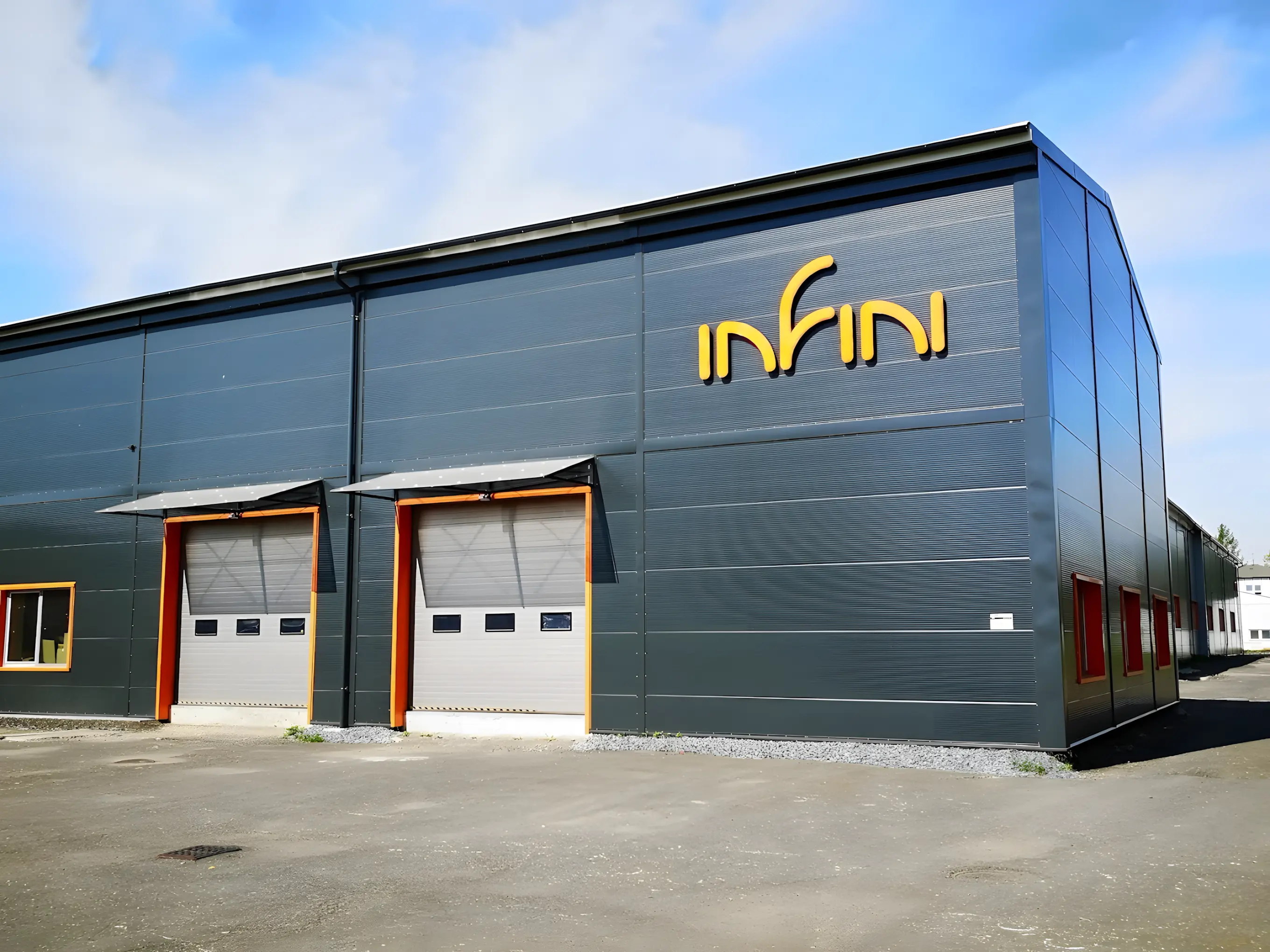 Moderní průmyslová hala společnosti Infini s tmavým kovovým opláštěním a žlutým logem na fasádě. Budova má dvě velká nakládací vrata s oranžovými rámy a několik oken po stranách. Před halou je zpevněná plocha a modrá obloha v pozadí dodává fotografii svěží vzhled.