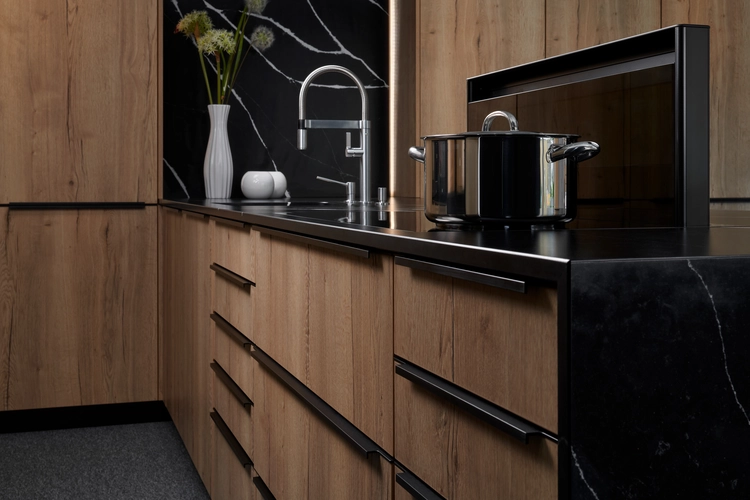 Moderní kuchyňská linka s dřevěnými předními plochami v teplém hnědém tónu, černou žulovou pracovní deskou a černým pozadím. Na lince je umístěn černý hrnec, stylová vodovodní baterie a minimalistické dekorativní nádoby v bílé barvě. Design kuchyně kombinuje přírodní dřevěné prvky s elegantními černými akcentními plochami.