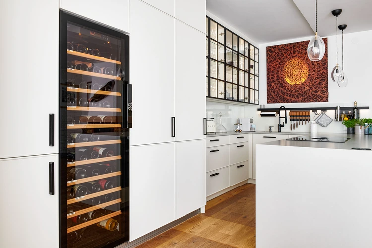 Luxusní kuchyně v moderním designu s bílými skříňkami a dřevěnou podlahou. Dominantou prostoru je vestavná vinotéka s prosklenými dvířky. Nad pracovní deskou je elegantní prosklená skříňka a na stěně visí umělecký obraz s oranžovými tóny.