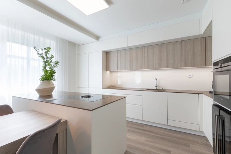 Minimalistická světlá kuchyně v bílé a dřevěné barevnosti. Prostorný interiér s kuchyňským ostrovem, vestavěnými spotřebiči, světlou pracovní deskou a velkou prosklenou stěnou s denním světlem. Zelená pokojová rostlina v designové váze dodává prostoru živost a přírodní prvek.