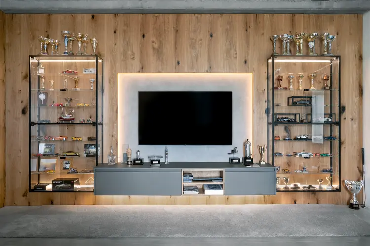 Luxusní obývací stěna s černou TV obrazovkou uprostřed, lemovaná dvěma prosklenými policovými jednotkami. Police jsou plné různých trofejí, pohárů a modelů aut. Dřevěné obložení stěny v teplém hnědém tónu vytváří elegantní pozadí. Tlumené osvětlení zvýrazňuje sbírku předmětů a vytváří útulnou atmosféru.