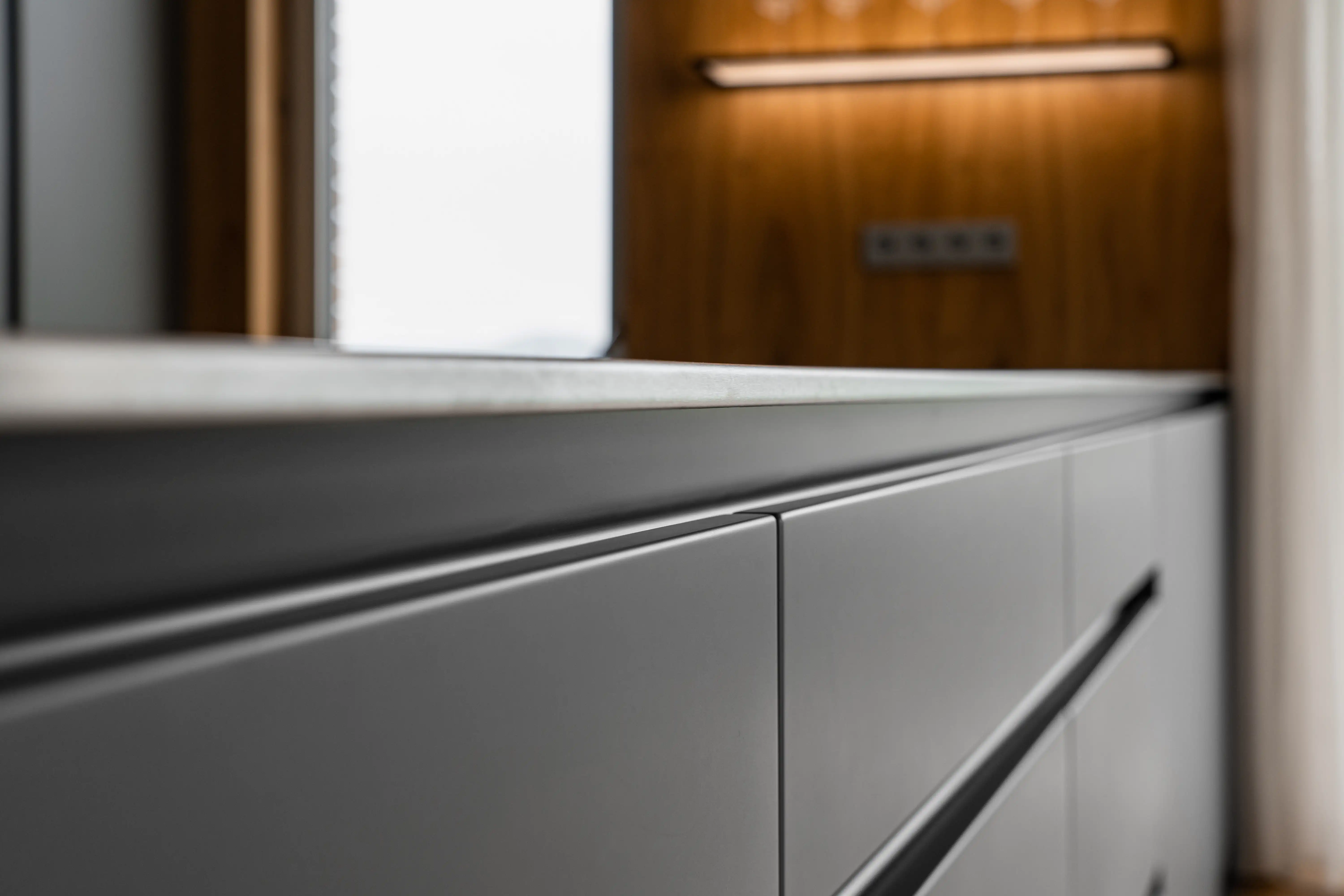Detail moderní kuchyňské linky s matně šedými bezúchytkovými skříňkami a světlou pracovní deskou. V pozadí je dřevěný obklad s integrovaným LED osvětlením, který dodává interiéru elegantní a útulný vzhled.
