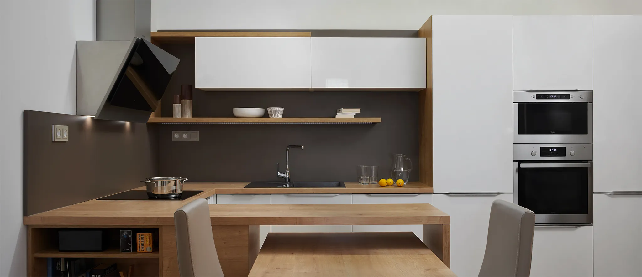 Minimalistická moderní kuchyně v šedé a bílé barvě. Designový interiér s vestavěnými spotřebiči, dřevěným jídelním stolem, otevřenými dřevěnými policemi a vestavěnou troubou. Čistý, prostorný design kombinující teplé dřevěné prvky s chladnými šedými plochami.