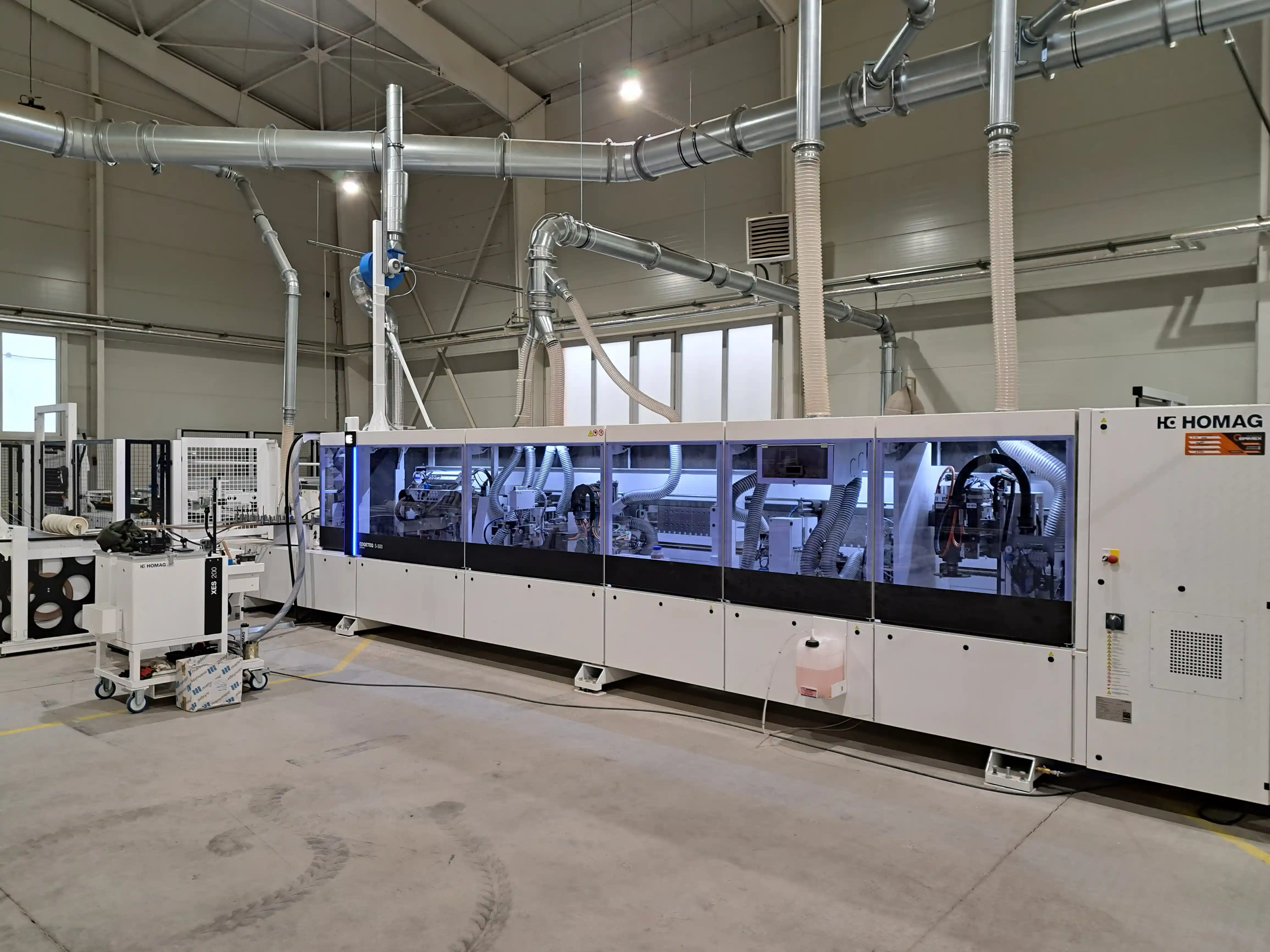 Průmyslový stroj HOMAG v moderní výrobní hale. Stroj je součástí automatizované linky na zpracování materiálů, obklopený kovovým potrubím pro odsávání prachu. Velká okna a světlé stěny poskytují dostatek přirozeného i umělého osvětlení, což zajišťuje efektivní pracovní prostředí.