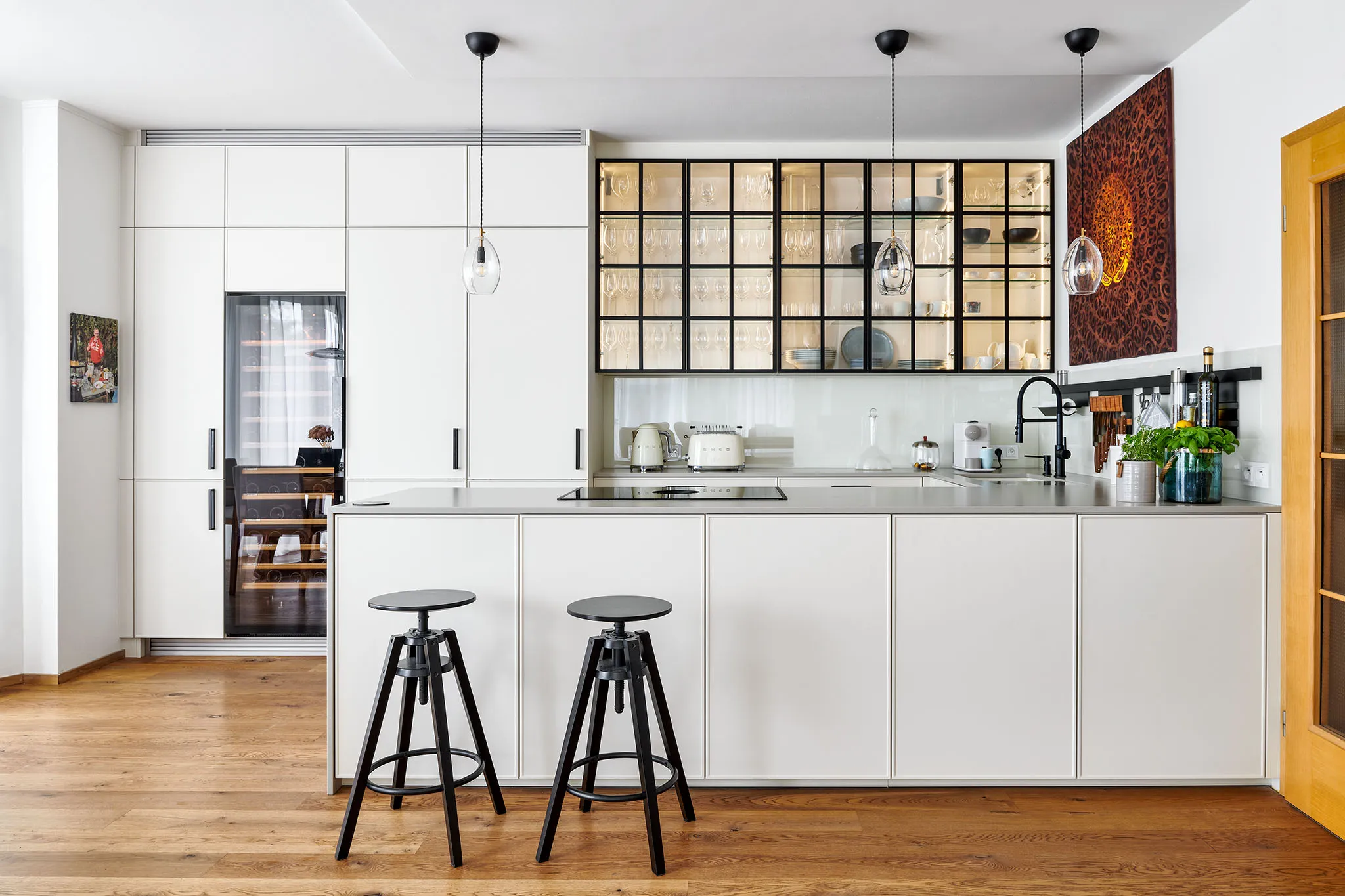 Světlá moderní kuchyň s bílými skříňkami a černými úchyty, vestavěnou vinotékou se skleněnými dveřmi, prosklenými horními vitrínami, kuchyňským ostrůvkem a závěsnými světly; na stěně je výrazný obraz v teplých barvách.