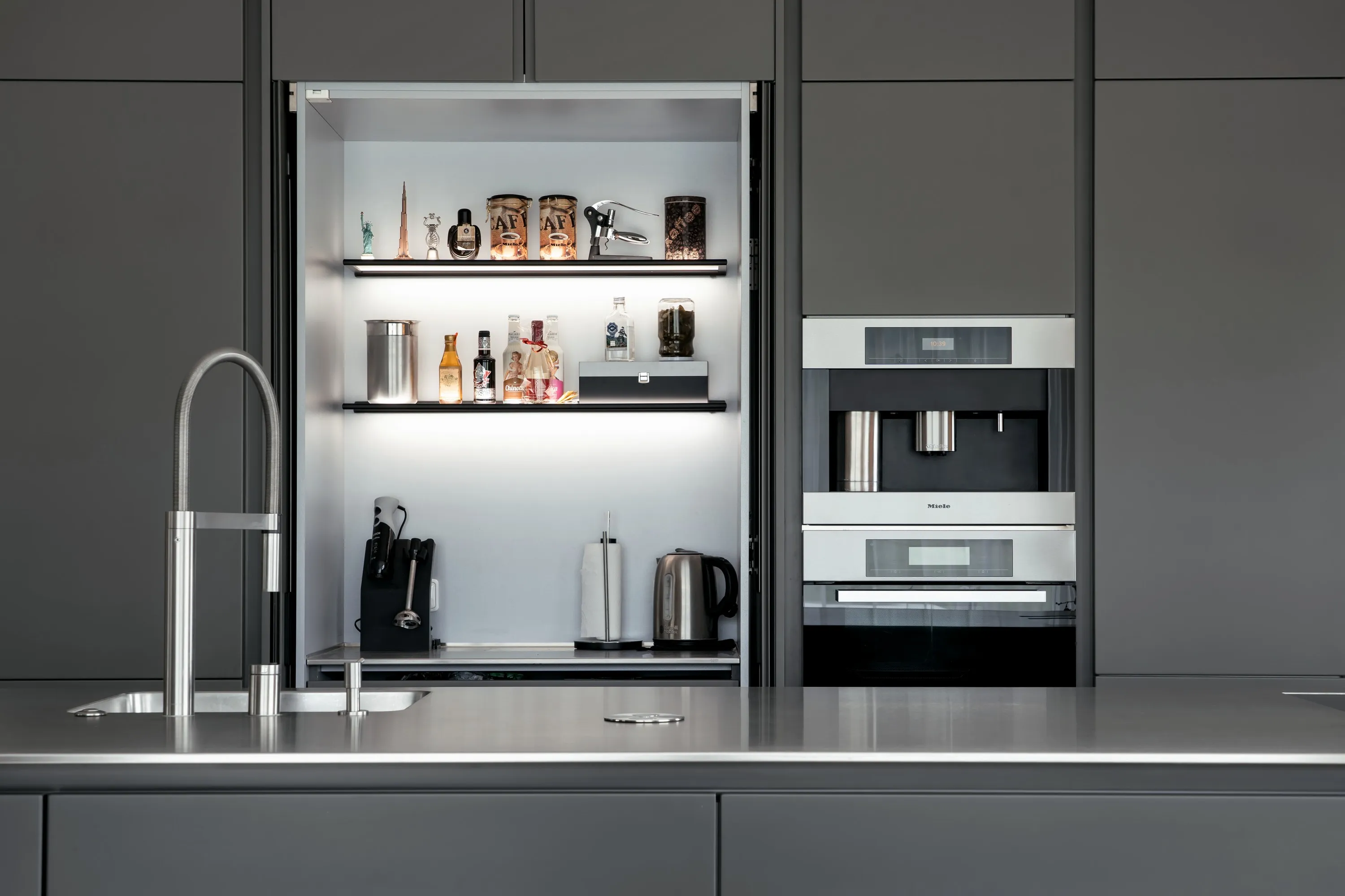 Moderní minimalistická šedá kuchyně s lesklými dvířky, nerezovým dřezem s designovou baterií, vestavěnými spotřebiči v černé a stříbrné barvě a praktickým úložným prostorem s osvětlenými policemi na drobné spotřebiče a dekorace