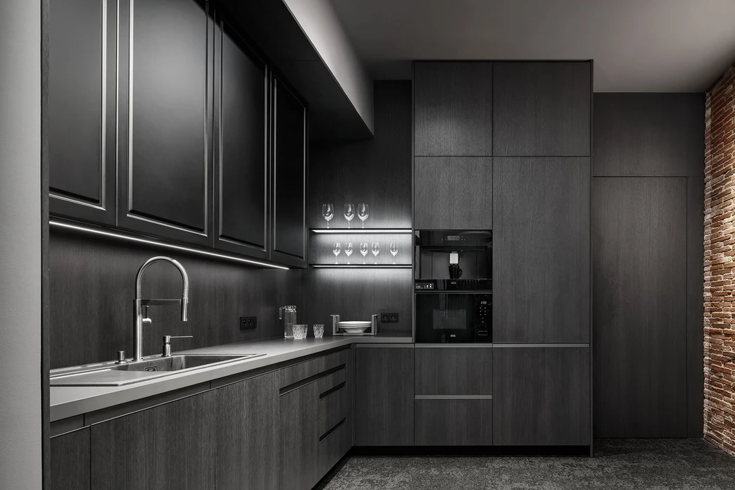 Moderní tmavá kuchyně v antracitovém provedení s LED podsvícením, vestavěnými spotřebiči, nerezovým dřezem, cihlovou stěnou, bezúchytkovým systémem otevírání a elegantní výstavkou na sklenice v minimalistickém designu v našem studiu INFINI v Říčanech
