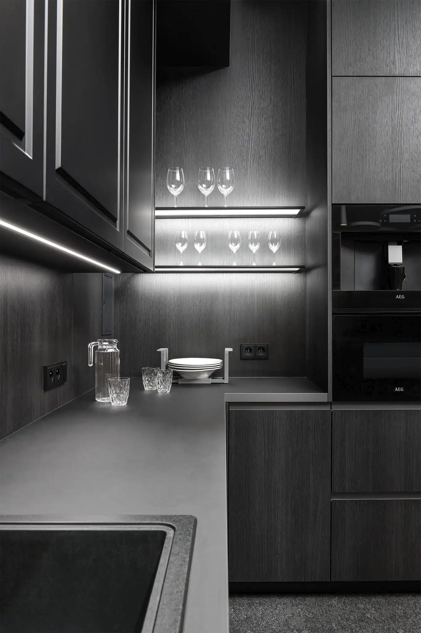 Detailní pohled na moderní tmavou kuchyň s podsvícenou pracovní plochou, kontrastním LED osvětlením pod skříňkami, elegantními tmavými povrchy a minimalistickým designem kuchyňské linky