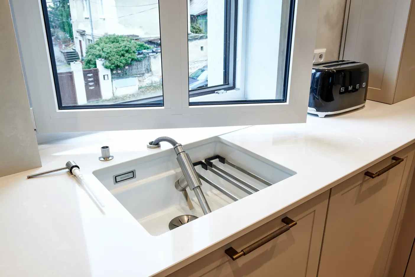 Stylový kuchyňský dřez s výsuvnou baterií umístěný pod oknem, ideální pro moderní kuchyně s důrazem na design a funkčnost.
