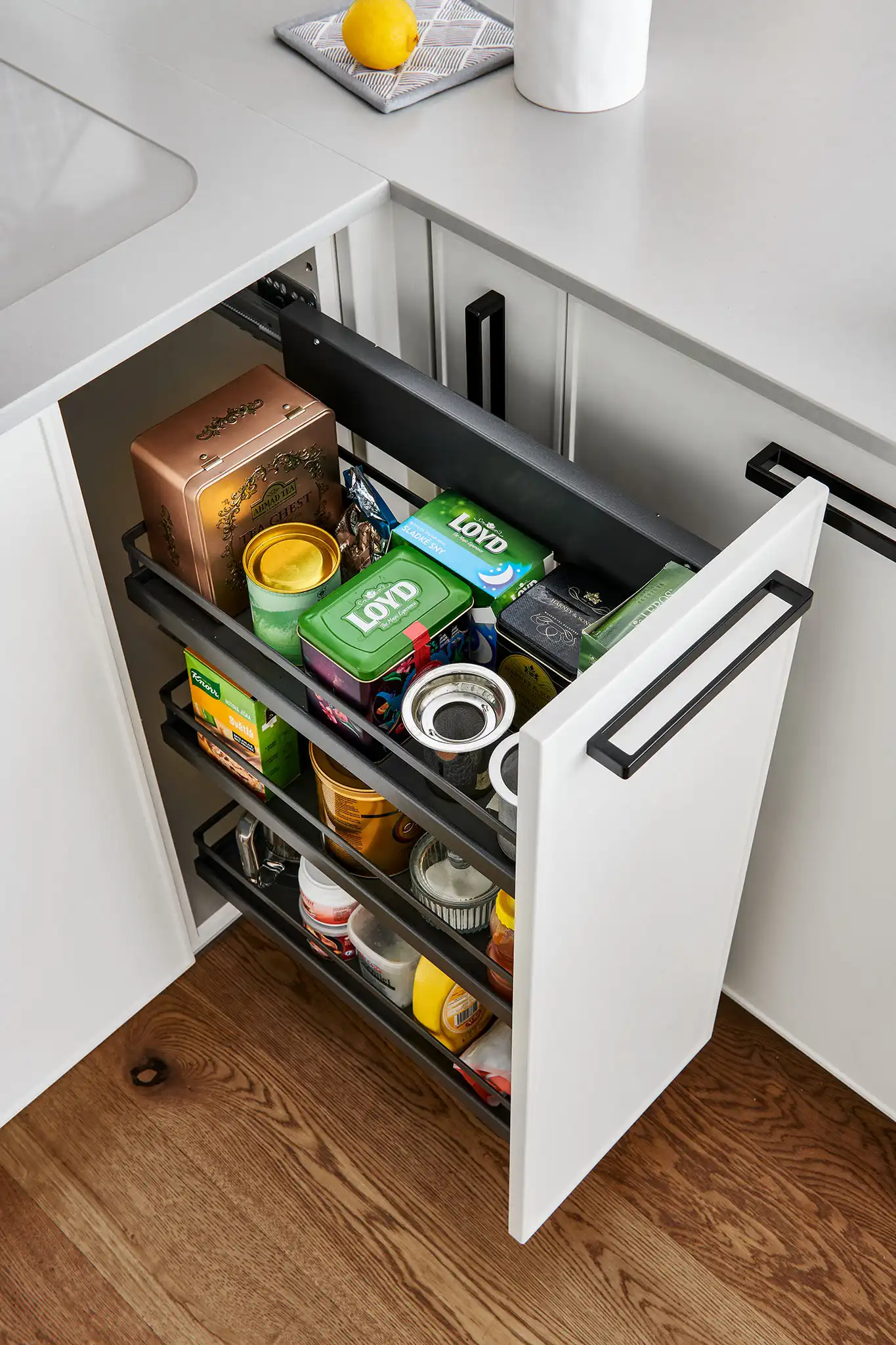 Široká výsuvná kuchyňská skříň s organizovaným úložným prostorem pro potraviny – plný výsuv s přehledným uspořádáním plechovek, sáčků a dóz v moderní bílé kuchyni.