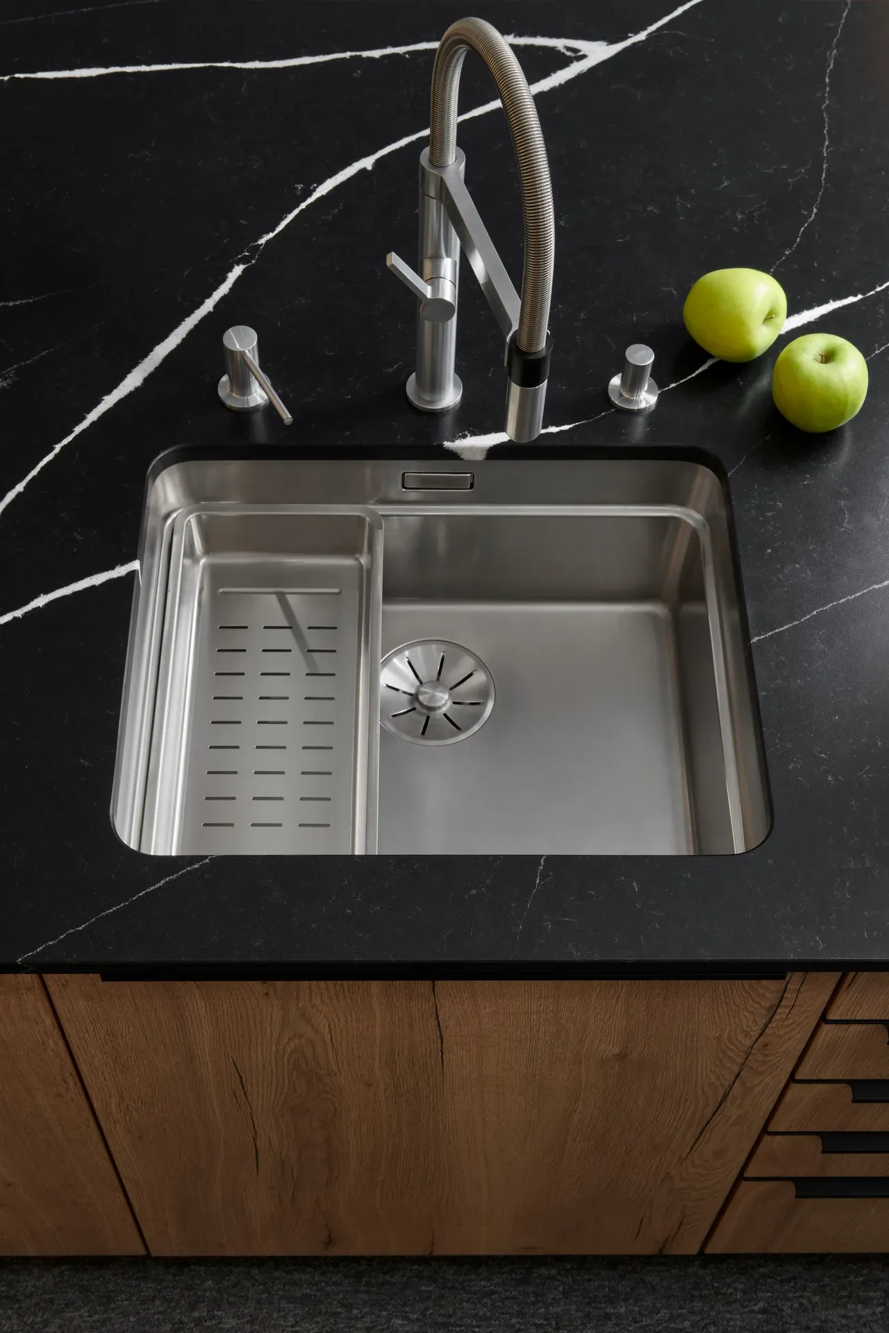 Nerezový kuchyňský dřez zapuštěný do černé pracovní desky s bílým žilkováním, doplněný moderní baterií a dávkovačem saponátu.