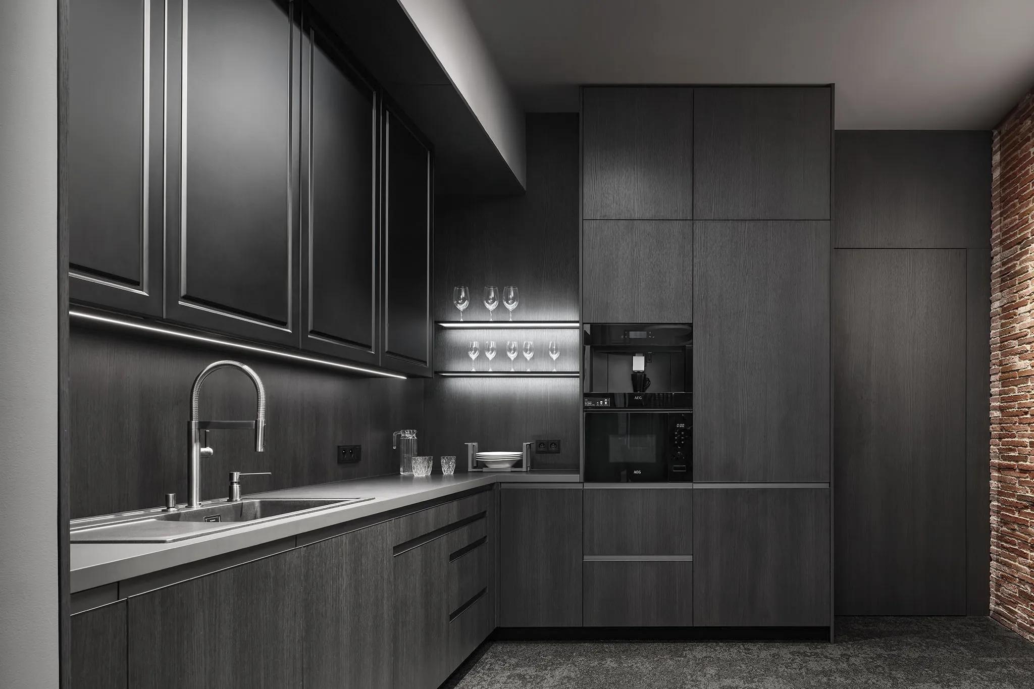 Moderní kuchyň v tmavém dekoru s nerezovým dřezem, vysokou baterií a vestavnými spotřebiči AEG, doplněná cihlovou stěnou.