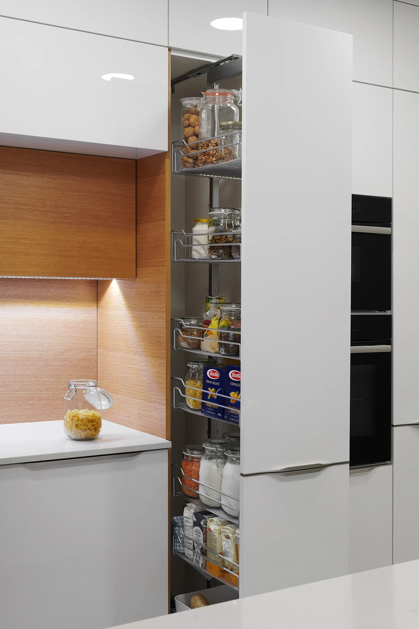 Moderní kuchyně s praktickým vysokým výsuvným systémem spížové skříňky s chromovými košíky plnými potravin, v kombinaci bílých a dubových dvířek s LED osvětlením a vestavěnými spotřebiči