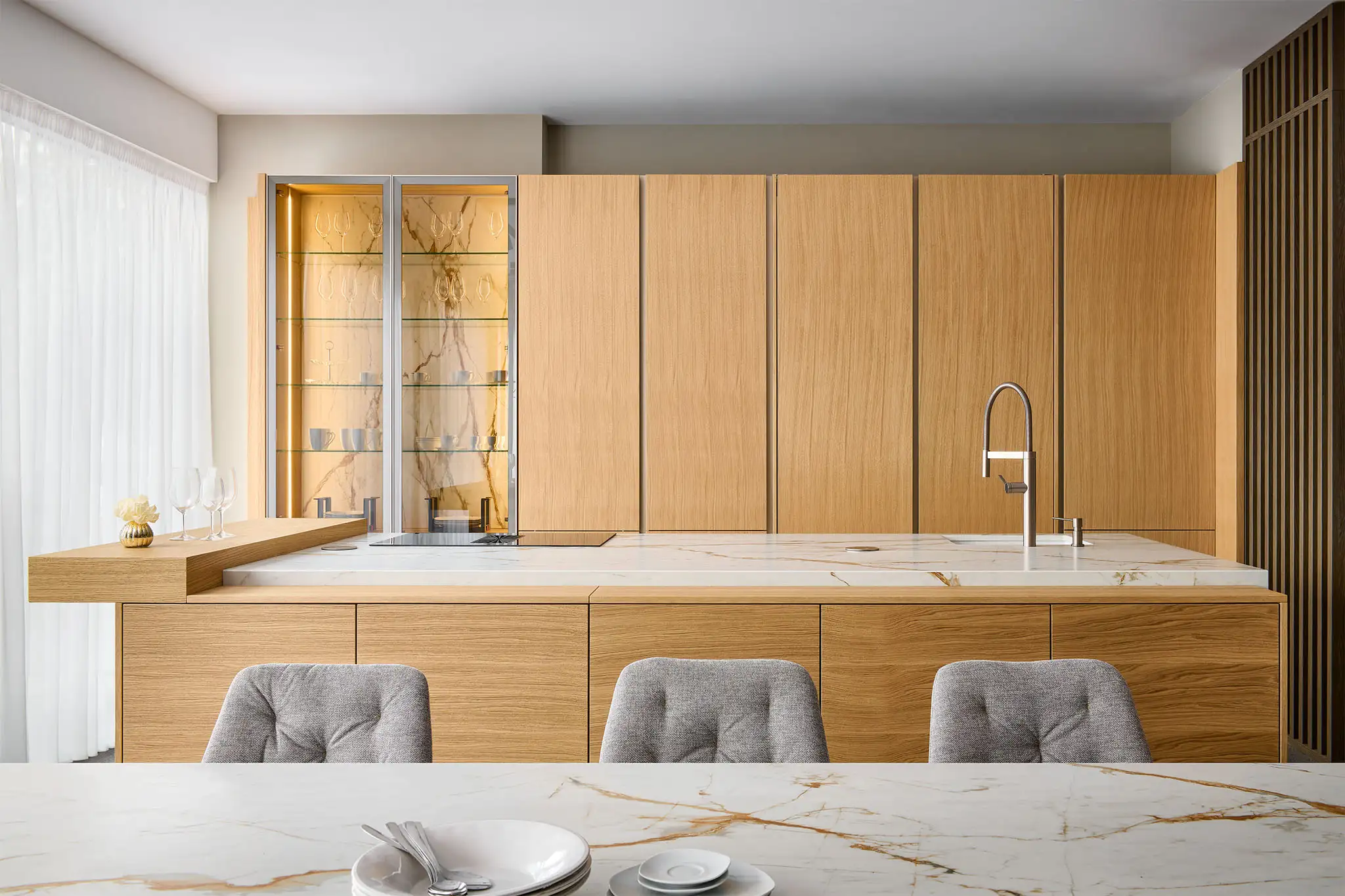 Minimalistická kuchyně s vestavnými spotřebiči ukrytými za vysokými dubovými skříňkami, mramorovou pracovní deskou, osvětlenou prosklеnou vitrinou a integrovaným dřezem pro čistý a elegantní vzhled
