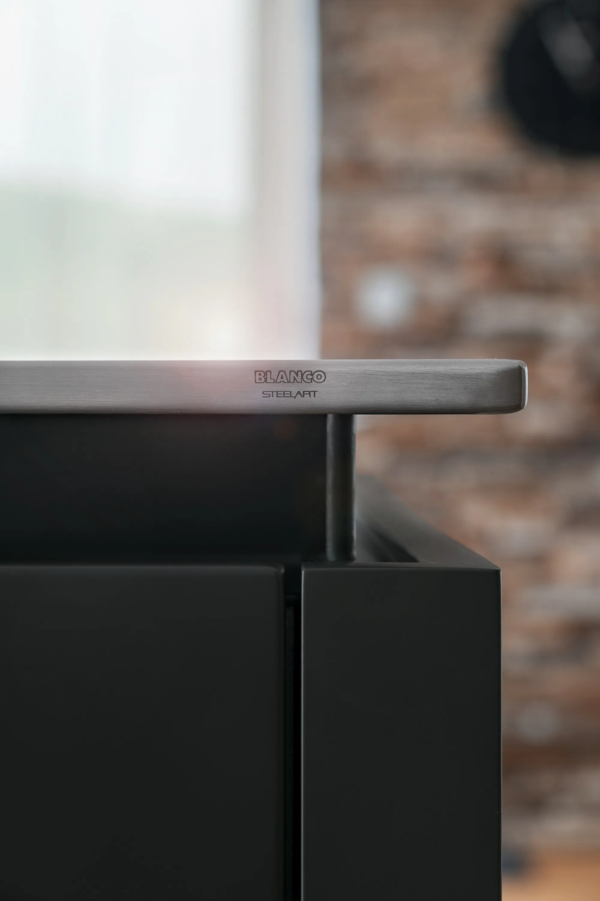 Detail nerezové hrany pracovní desky BLANCO STEELART s matným povrchem, přesahující přes černý kuchyňský nábytek v moderní kuchyni