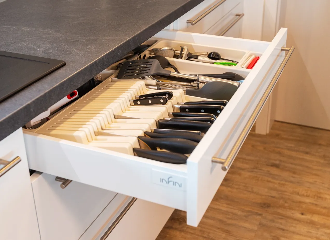Výsuvná kuchyňská zásuvka s přehledným organizérem na nože a kuchyňské náčiní, umístěná pod pracovní deskou v moderní bílé kuchyni s kovovým madlem.