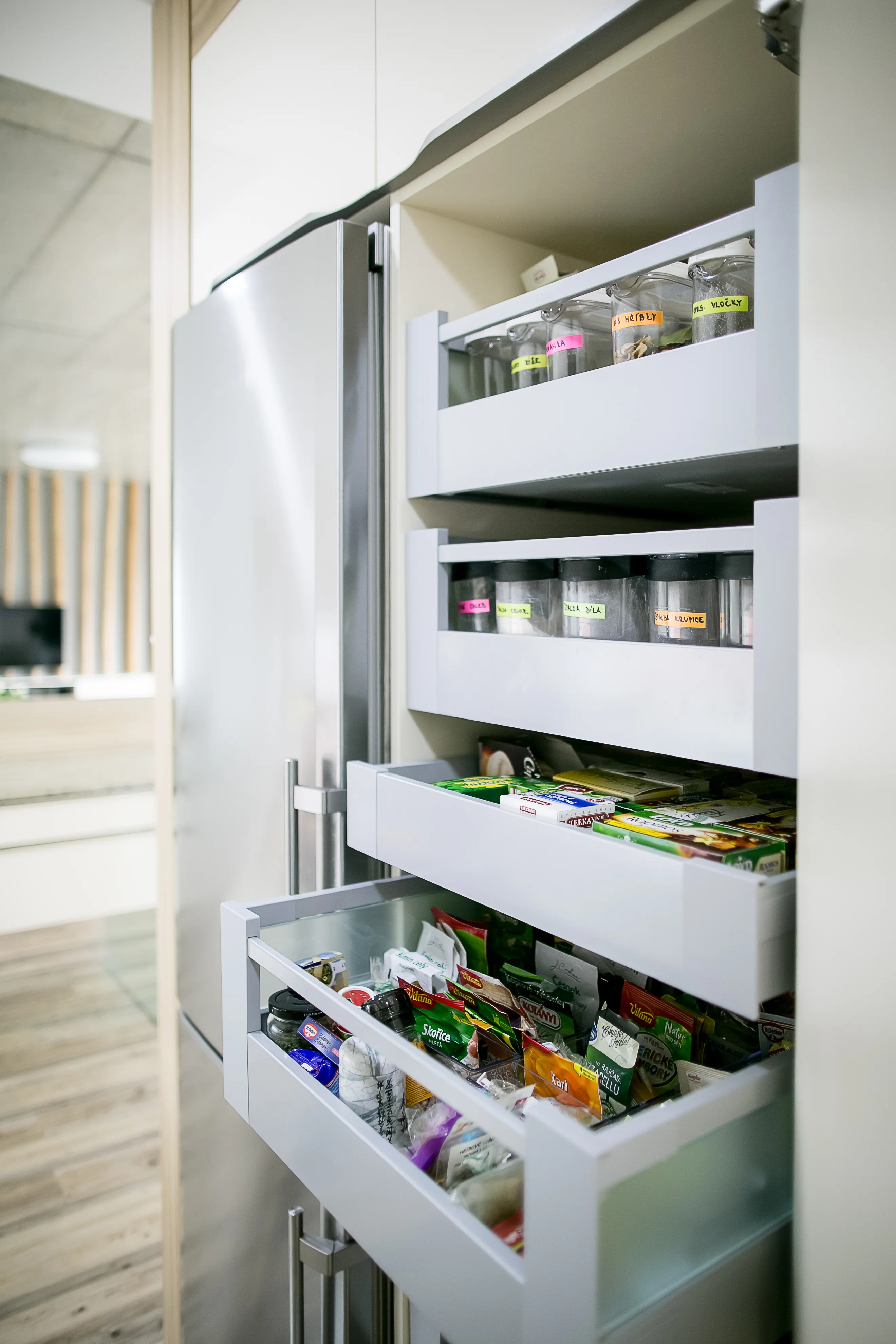 Vysoká výsuvná potravinová skříň s několika zásuvkami, přehledně uspořádanými kořenkami a balenými potravinami, umístěná v moderní kuchyni vedle vestavné lednice.
