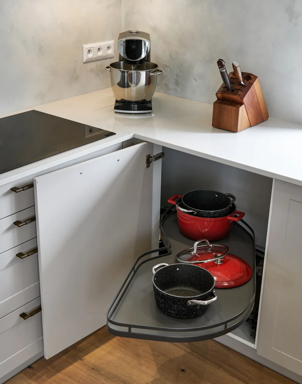 Rohová kuchyňská skříň s výsuvným otočným mechanismem na hrnce, otevřená dvířka pod pracovní deskou v moderní bílé kuchyni s indukční varnou deskou a spotřebiči na lince.
