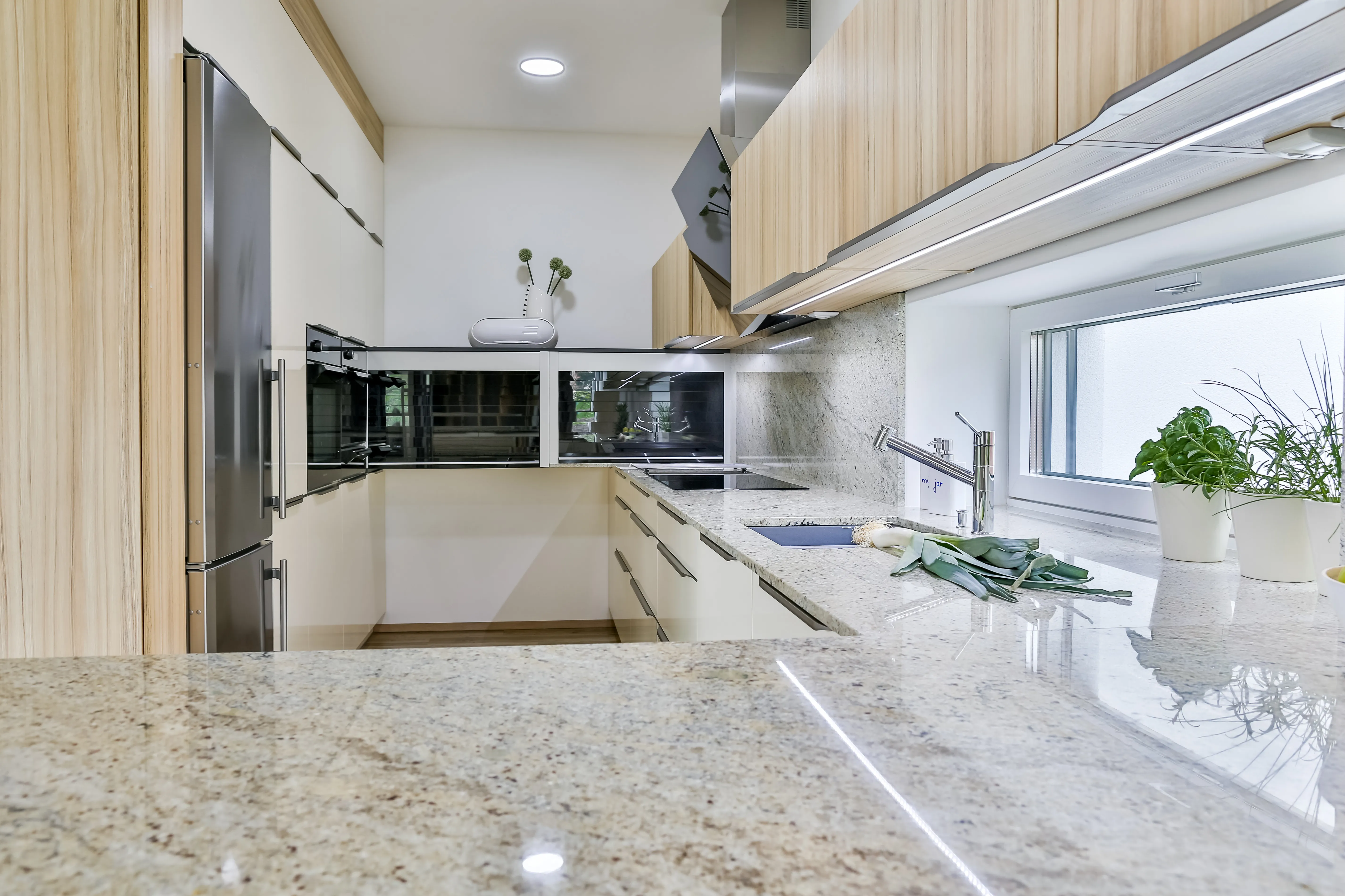 Detail moderní kuchyně na míru s kamennou pracovní deskou, dřevěnými horními skříňkami, bílými spodními zásuvkami, integrovanými spotřebiči, nerezovou lednicí a osvětlenou pracovní plochou u okna