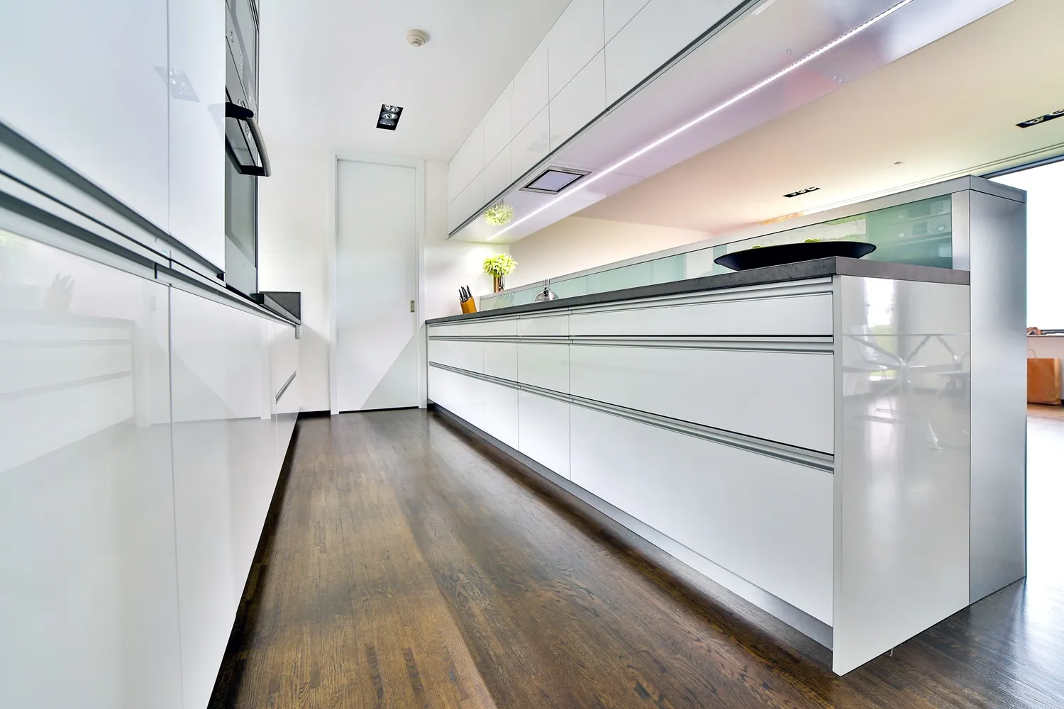 Minimalistická bílá kuchyně s bezúchytkovými skříňkami v lesku, dlouhou pracovní deskou v tmavém odstínu, podsvícenými horními skříňkami a tmavou dřevěnou podlahou, focená z nízké perspektivy podél linky.