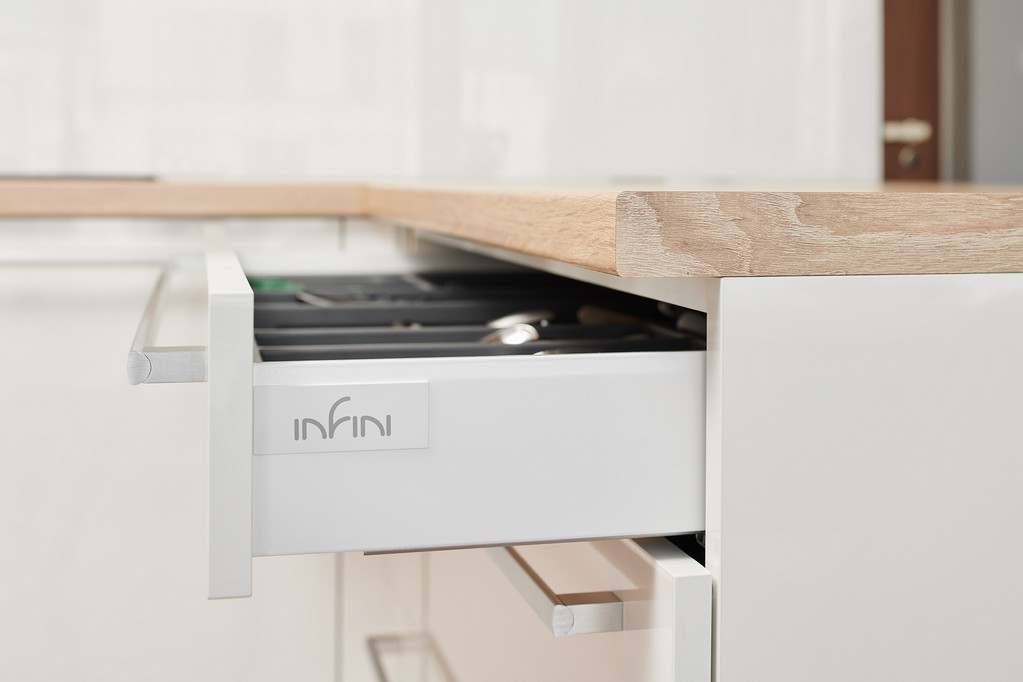 Detail bílé kuchyně INFINI s dřevěnou pracovní deskou, otevřeným výsuvem a čistým napojením materiálů.